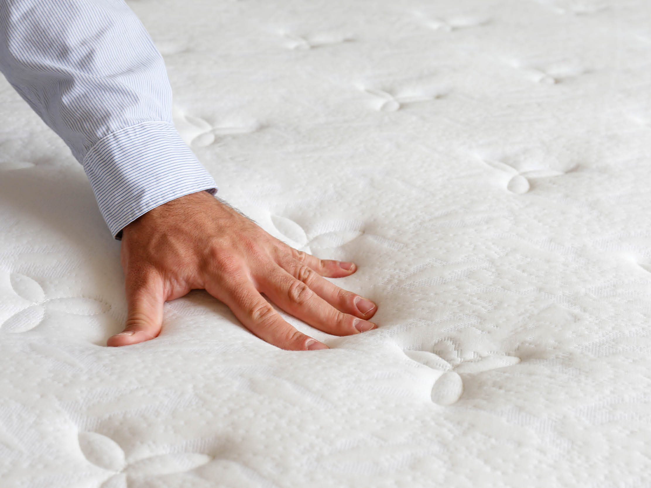 Cropped shot of man testing white orthopedic matress on firmness. Male pressing hypoallergenic foam mattresses surface to check its softness. Close up, copy space, top view, background.
