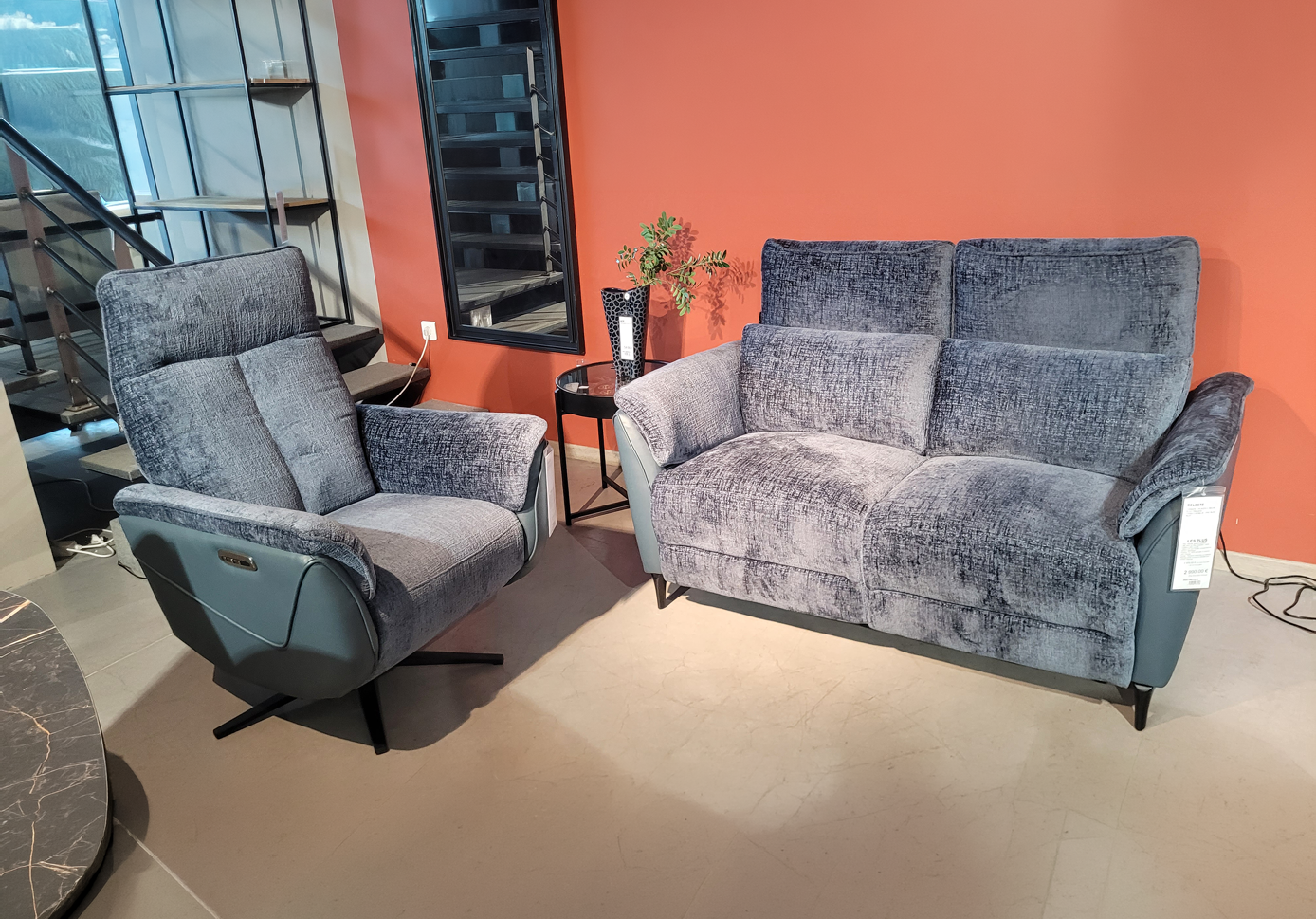 Un fauteuil et un canap gris en tissu moelleux sont dispos s dans un salon avec un mur orange et une petite table noire avec un vase et une plante.  Description g n r e par IA