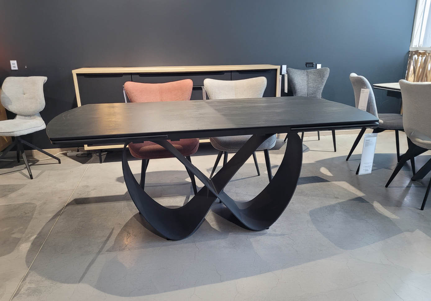 Table noire moderne avec un plateau rectangulaire et un pied sculptural en forme de ruban entrelac , entour e de plusieurs chaises dans un espace int rieur au sol en b ton. Description g n r e par IA