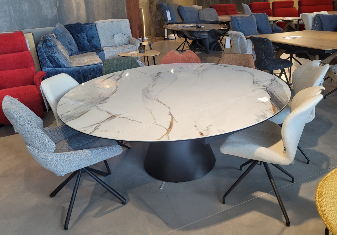 Une grande table ronde avec un plateau en marbre blanc et des veines dor es est entour e de six chaises rembourr es dans un showroom de meubles avec plusieurs canap s et tables en arri re-plan. Description g n r e par IA