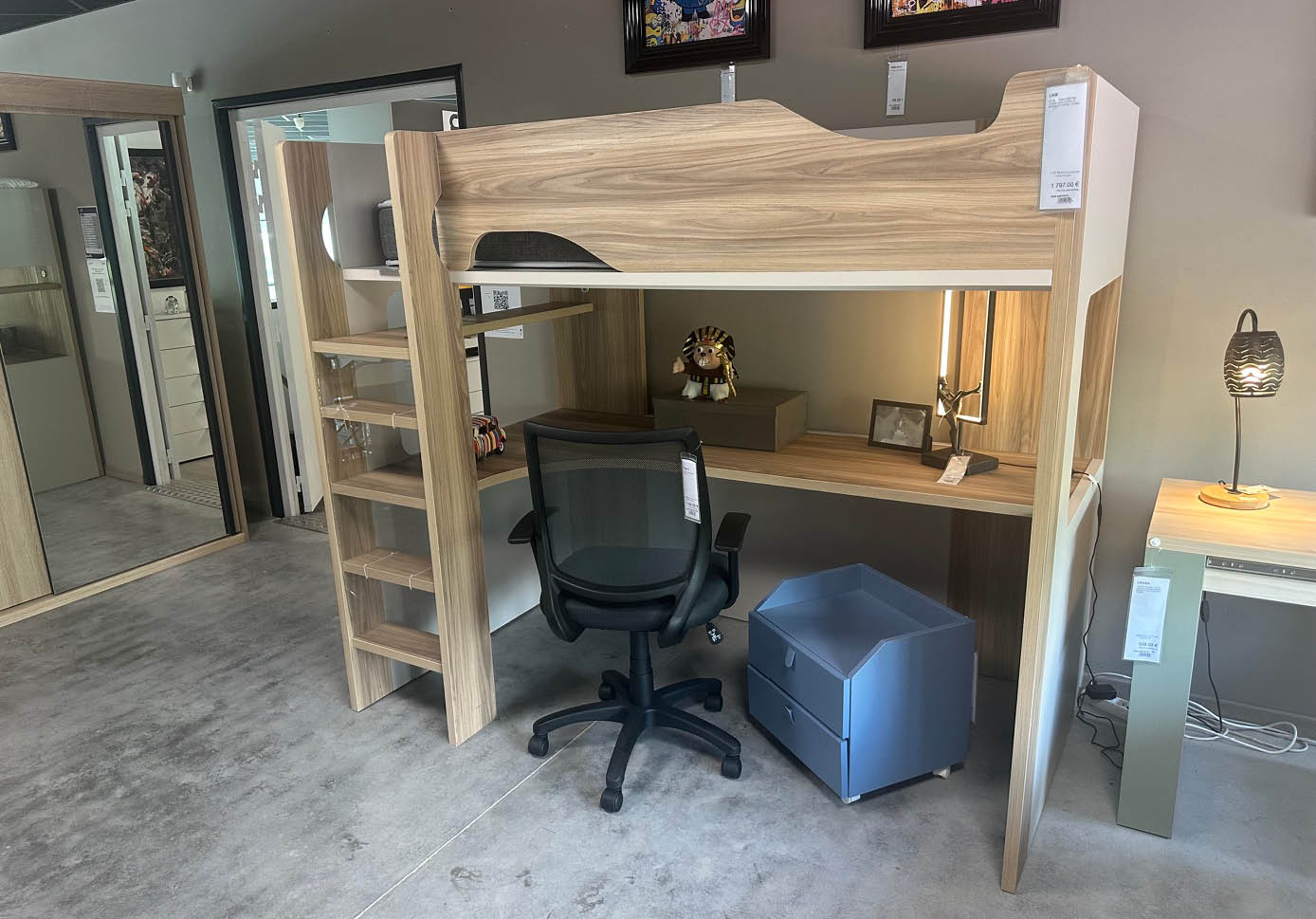 Un bureau en bois clair avec une chaise de bureau noire, un petit meuble bleu  roulettes, et une lampe pos e sur une autre table   c t  dans une pi ce au sol en b ton.  Description g n r e par IA